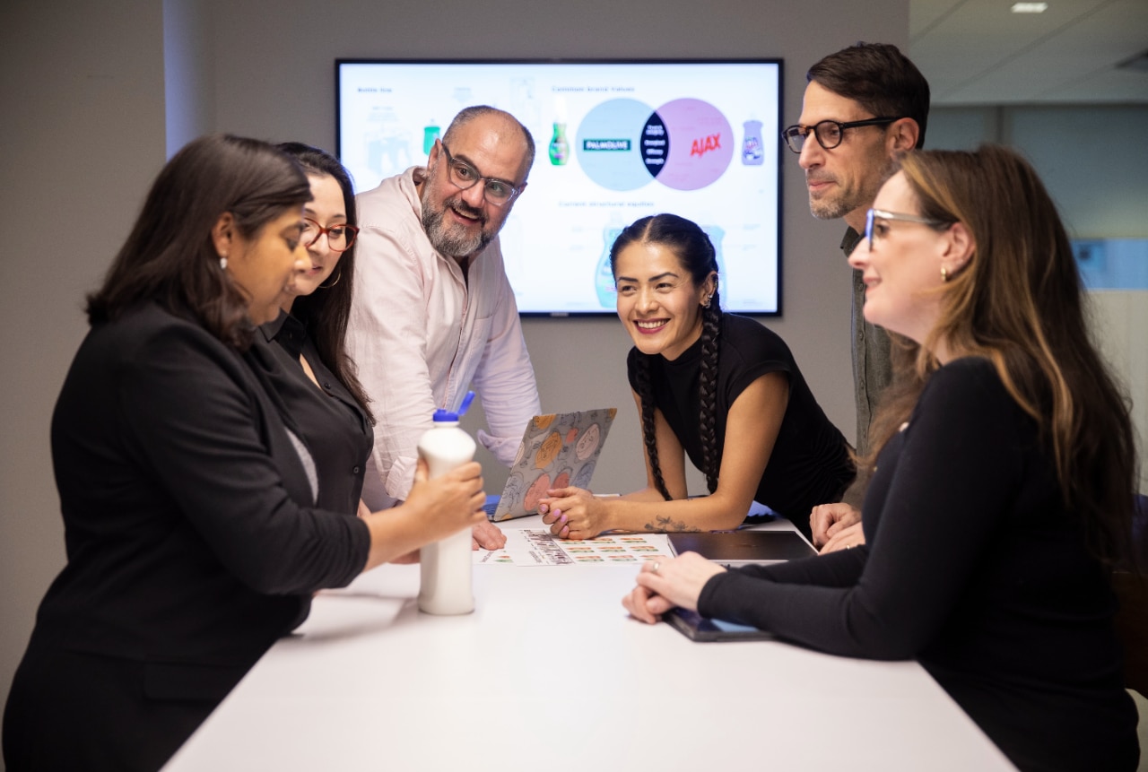 Seis empleados de Colgate-Palmolive en una sala de reuniones analizando un concepto de producto, con una diapositiva de presentación en una pantalla detrás de ellos.